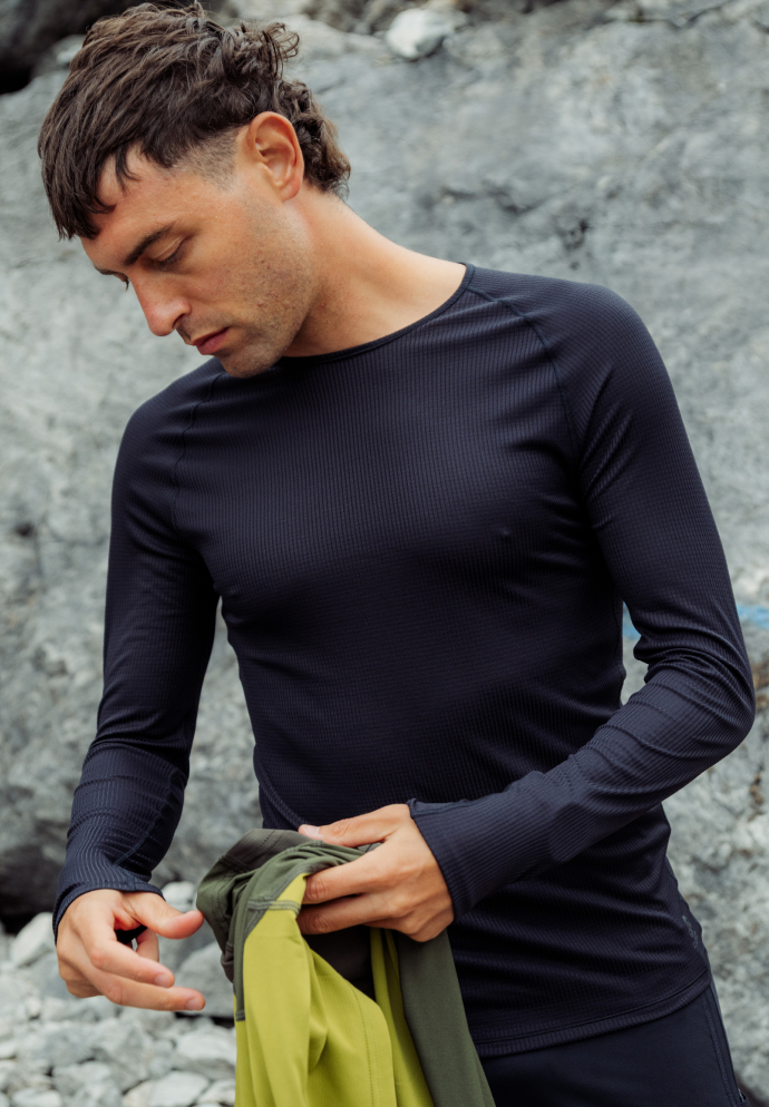 Man in a dark ribbed long-sleeve light base layer top stands by grey rocks, holding a green and yellow garment while looking down.
