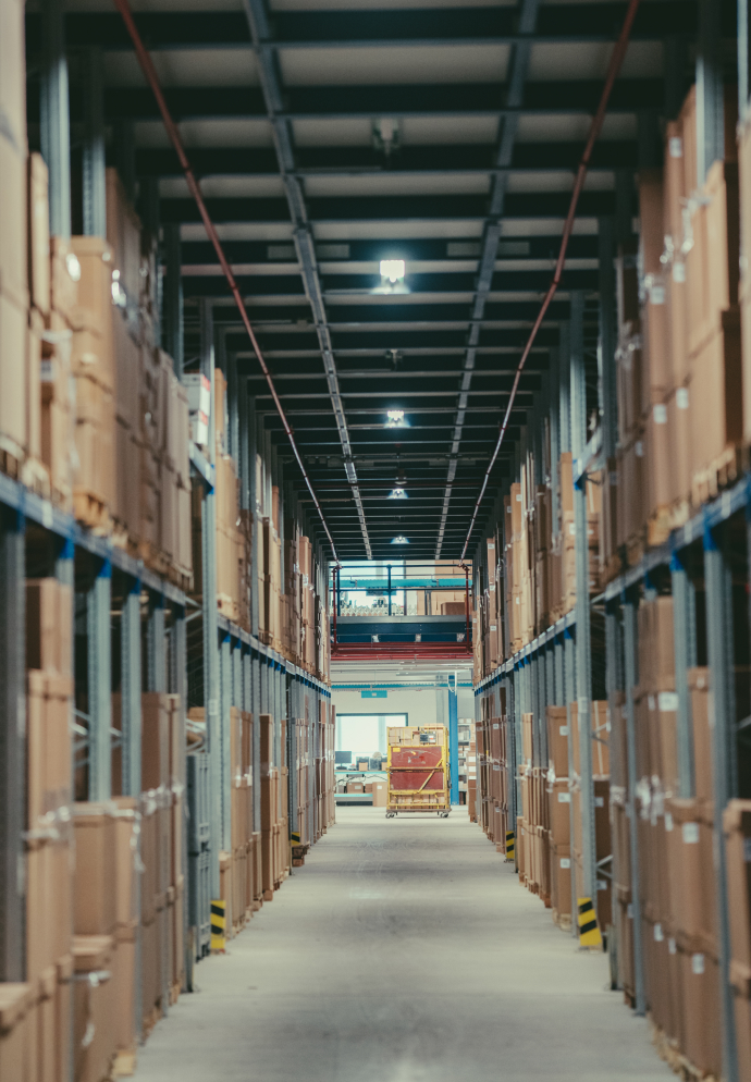 Long warehouse aisle flanked by tall metal shelves packed with brown cardboard boxes, leading to a yellow cart at the end.