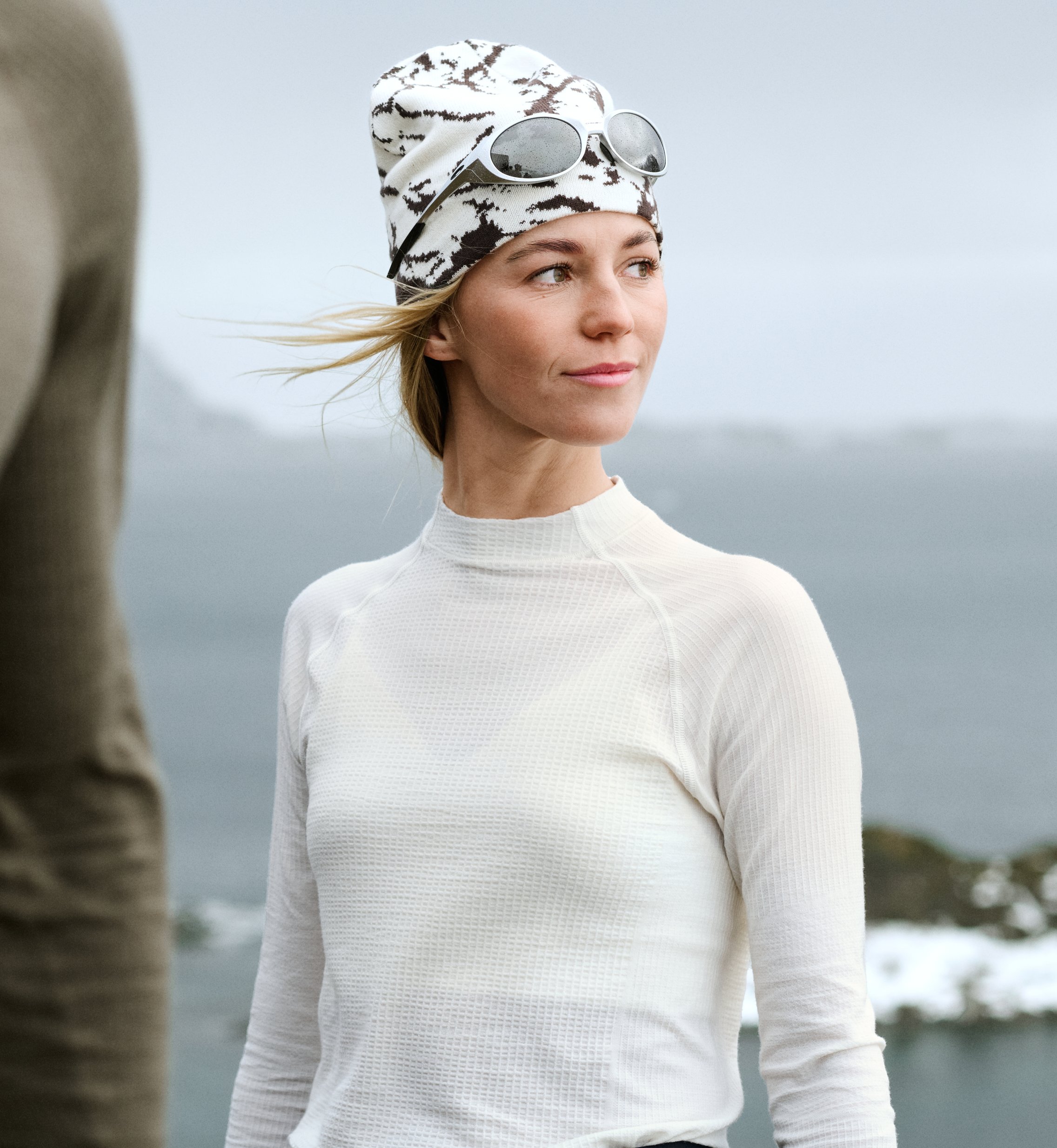 Donna con maglia termica bianca e berretto fantasia con occhiali da sole guarda verso destra. Persona sfocata in primo piano, acqua sullo sfondo.