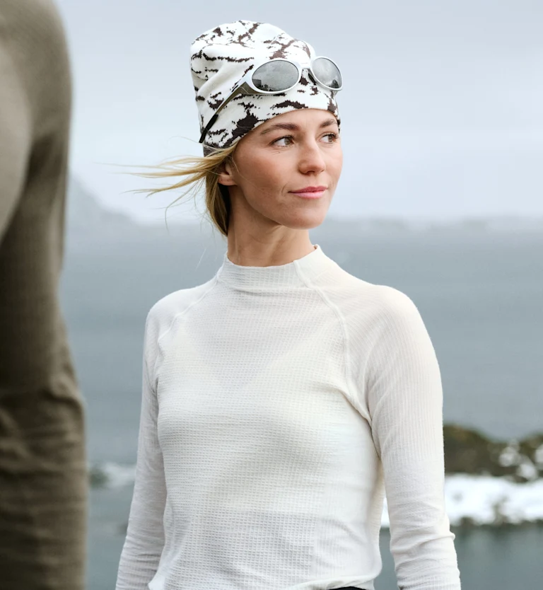 Donna con maglia termica bianca e berretto fantasia con occhiali da sole guarda verso destra. Persona sfocata in primo piano, acqua sullo sfondo.