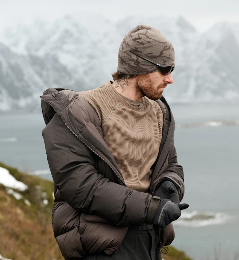 Bearded man in brown puffer jacket, beanie, and sunglasses adjusts gloves against a backdrop of snowy mountains and water.