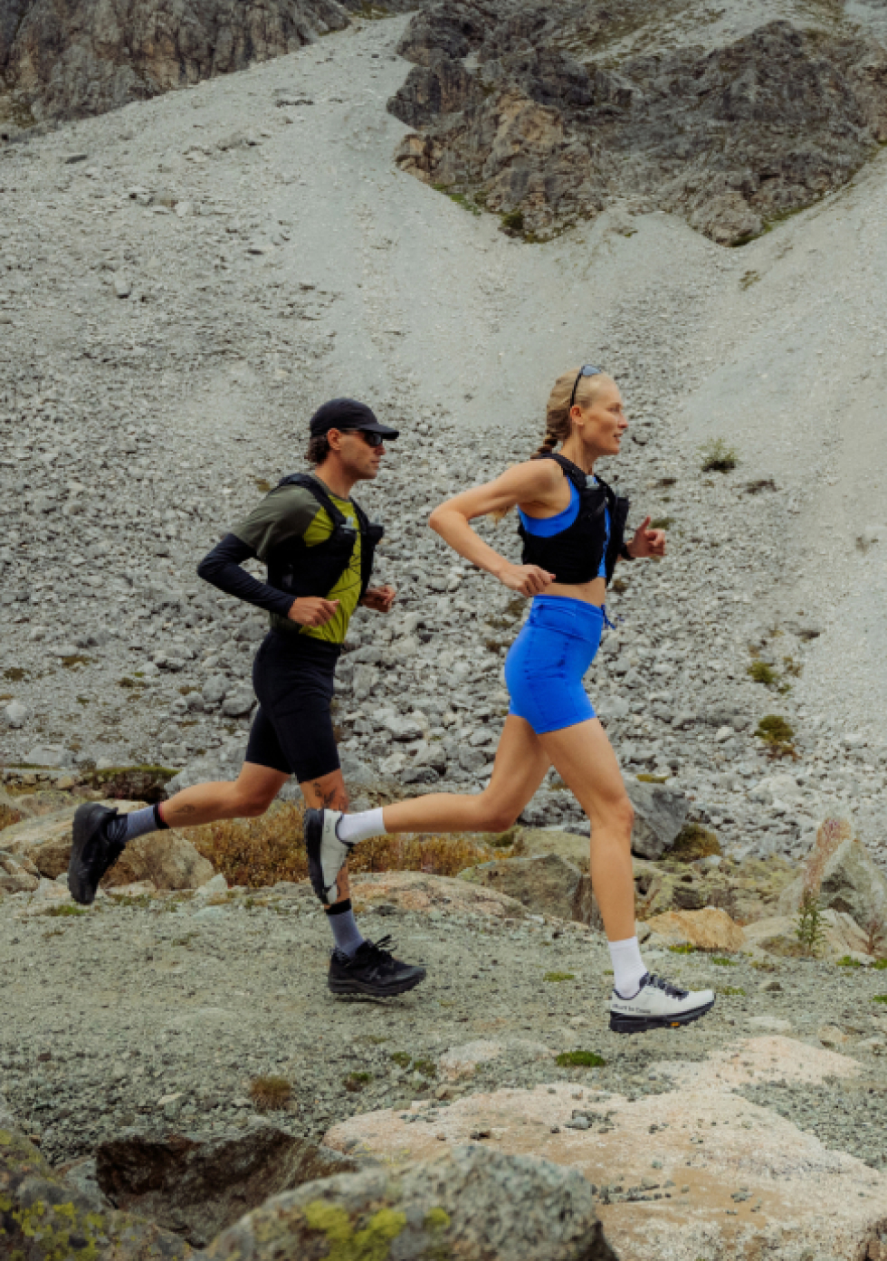 Een man in het groen en een vrouw in het blauw die trailrunnen op een rotsachtig bergpad, beiden met een zwart hydratatievest.