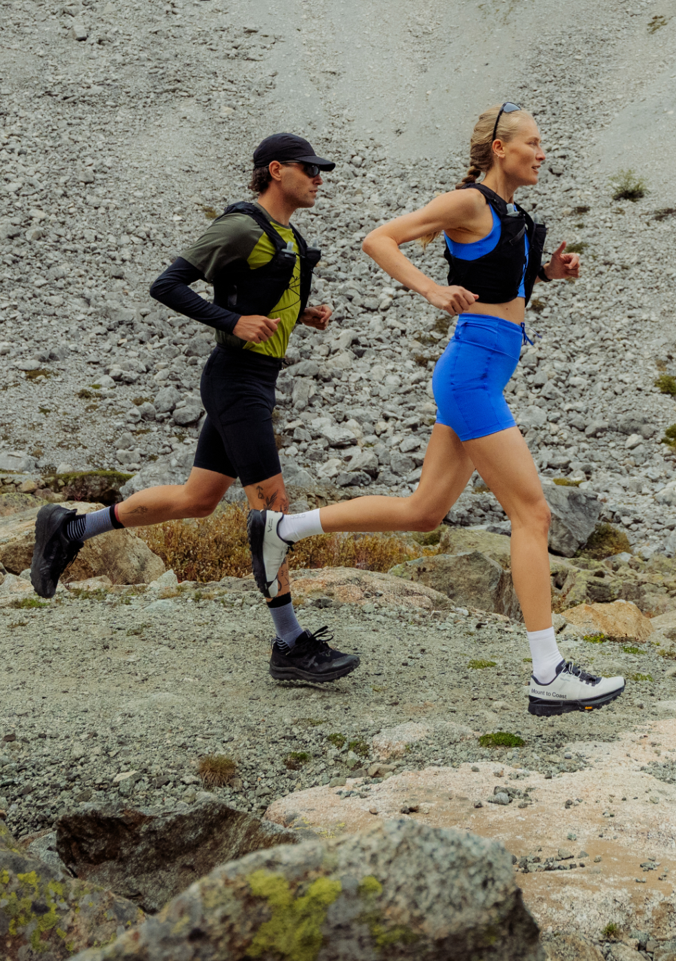 A man in green and a woman in blue trail running on a rocky mountain path, both wearing black hydration vests.