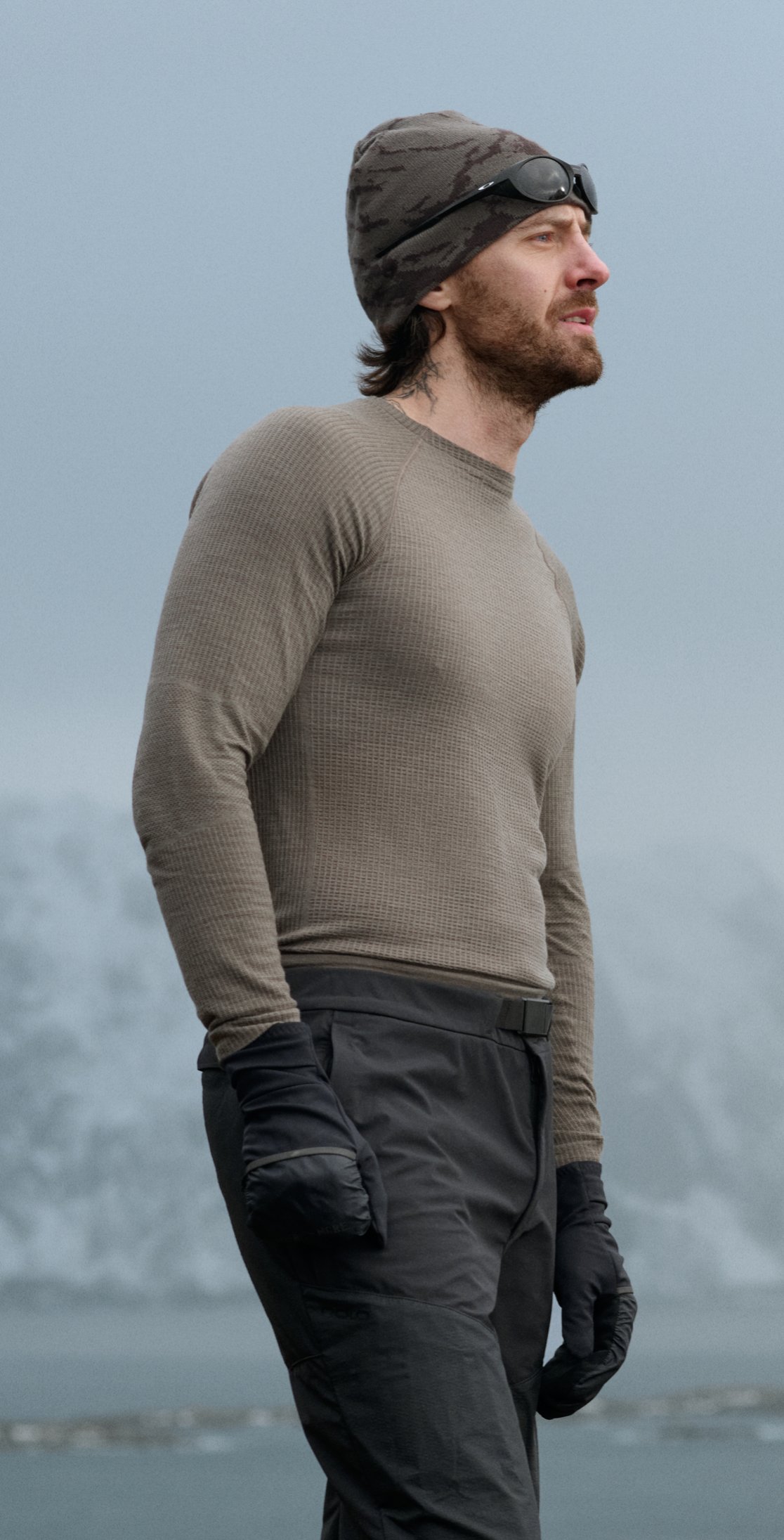 Homme barbu en haut thermique kaki, pantalon foncé, gants noirs et bonnet camouflage avec lunettes de soleil, regardant vers la droite en extérieur.