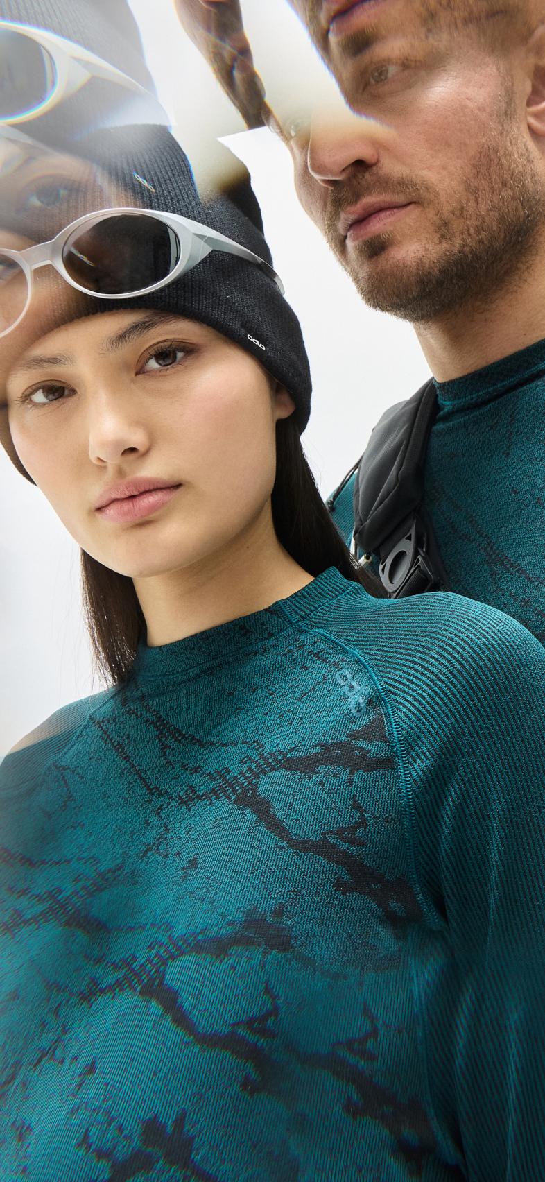 Woman in reflective orange ski goggles and black top stands by a man in a teal patterned shirt holding a black backpack.