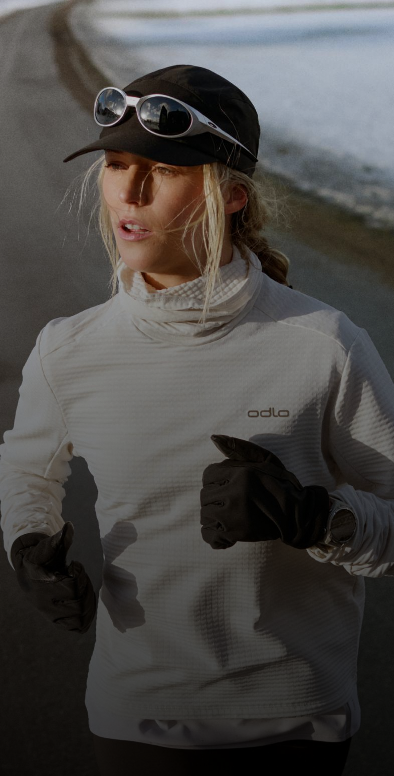 Woman running on a road in a snowy landscape, wearing a white Odlo running mid layer, black cap, sunglasses, and black gloves.