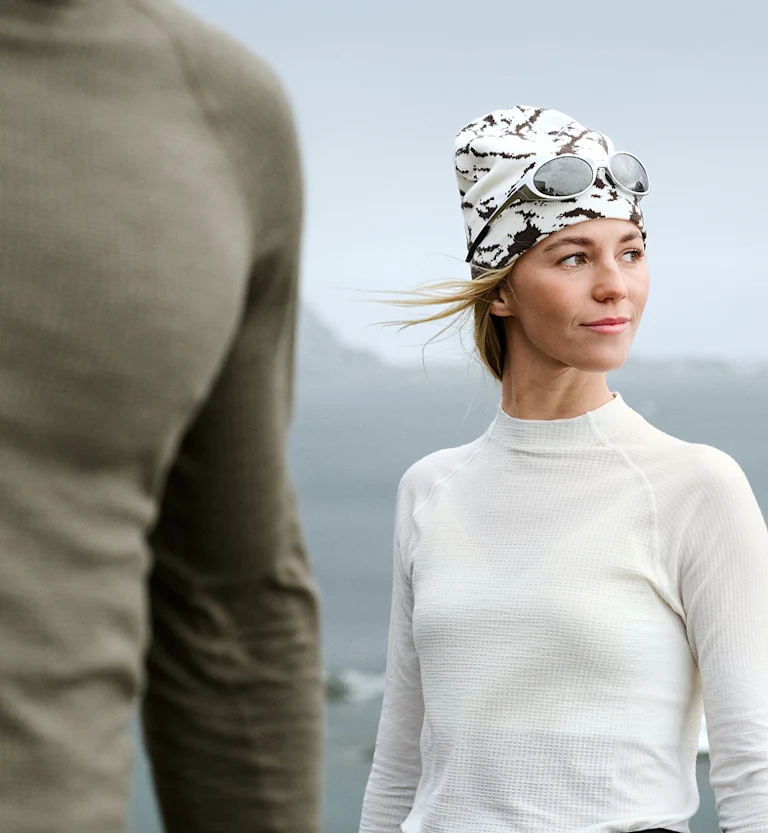 Woman in white thermal top and beanie looks right. Blurred person in green foreground. Overcast coastal background.