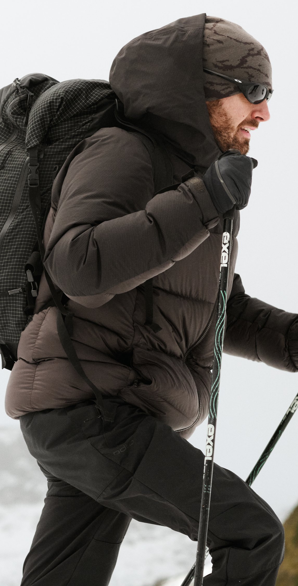 Een man met een baard, in een donkere gewatteerde jas en met een zonnebril, wandelt met een grote rugzak en trekkingstokken door de sneeuw.