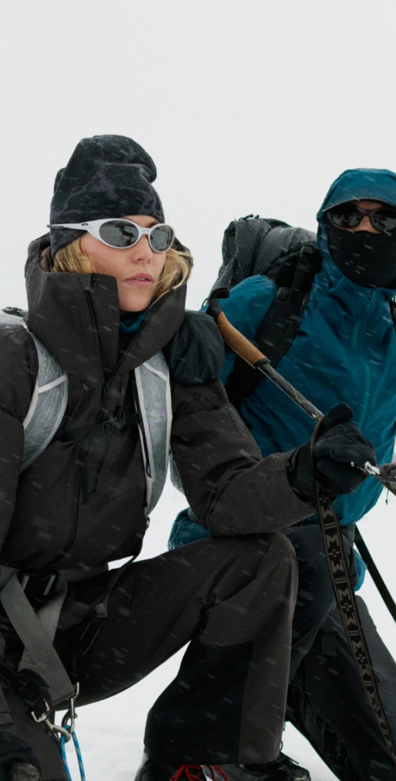 Donna in ginocchio e persona in abbigliamento blu che indica con un bastoncino da sci, entrambe con abbigliamento invernale, insieme a un husky in un paesaggio montano innevato.