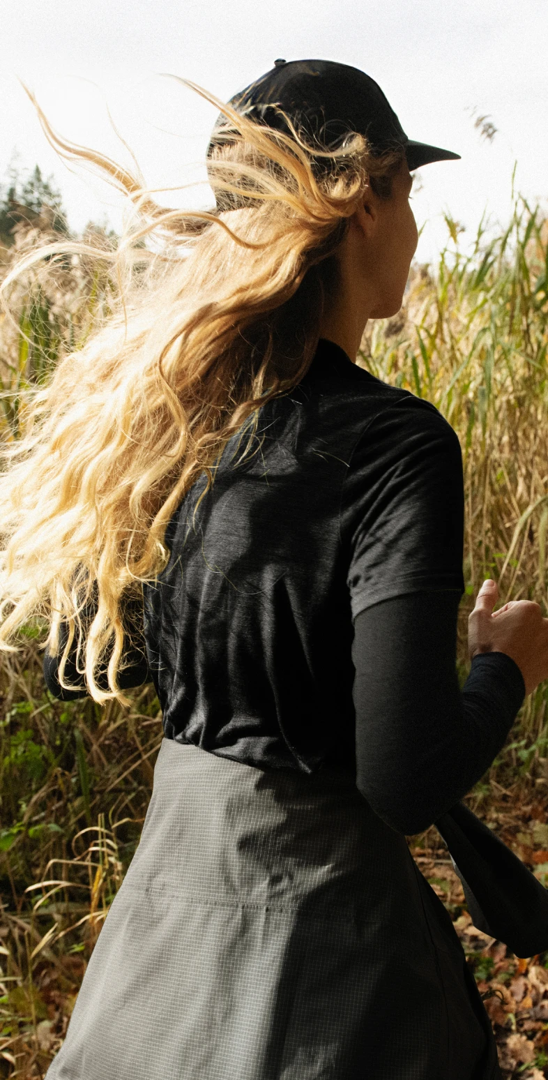 Blonde vrouw in een zwarte pet en shirt die rent op een pad door hoog droog gras, haar lange haar wapperend in de wind.