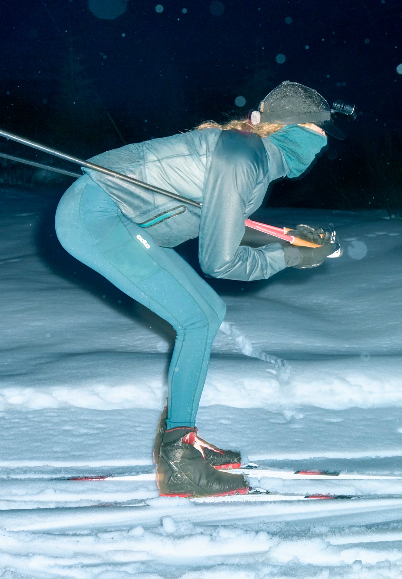 Skieur accroupi en tenue turquoise pratiquant le ski de fond de nuit sous la neige qui tombe, portant une veste imperméable double couche, une lampe frontale et un cache-visage.