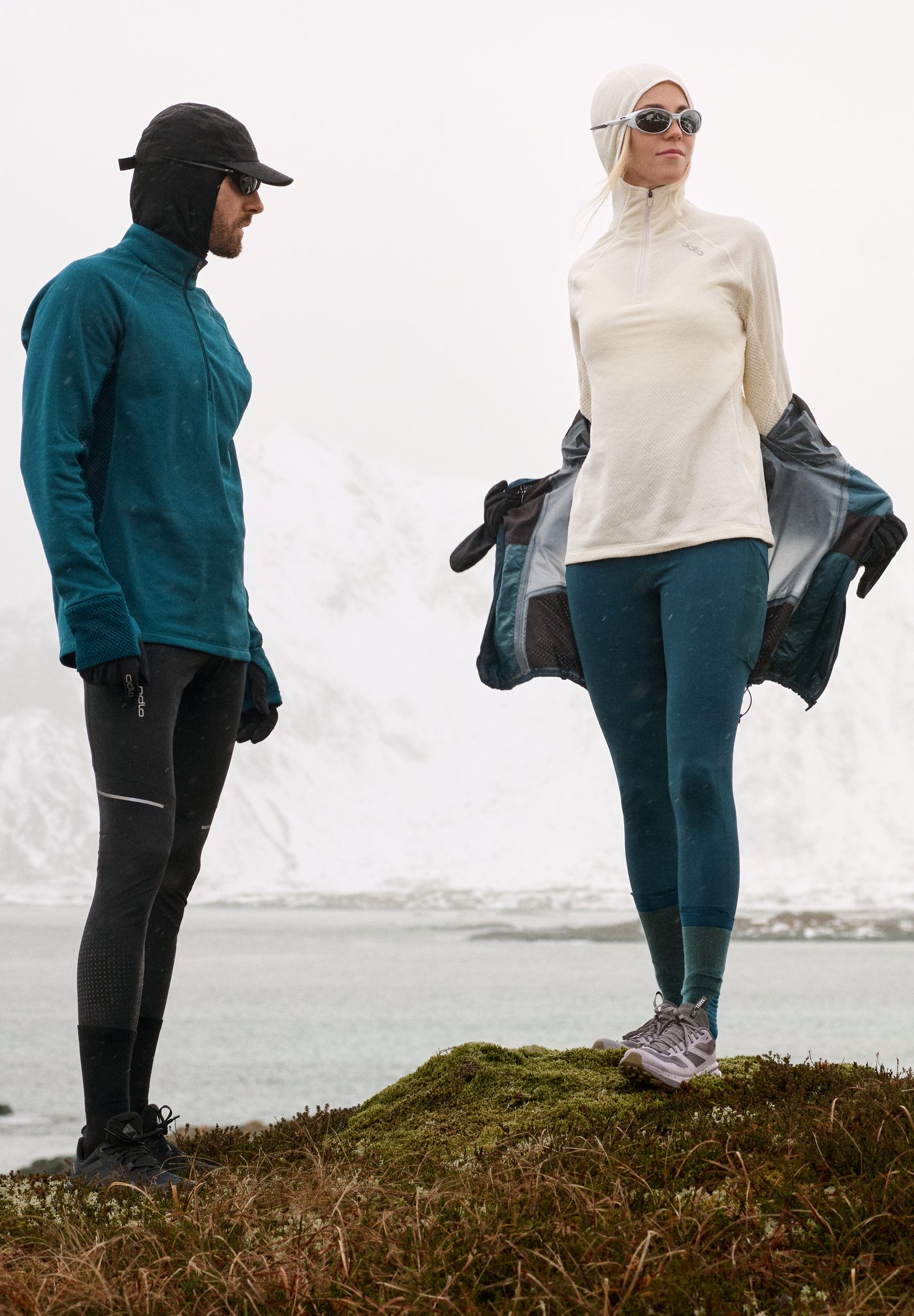 Two hikers in winter gear stand on mossy ground by a snowy fjord; woman removes a jacket, both wear hats, sunglasses, and leggings.