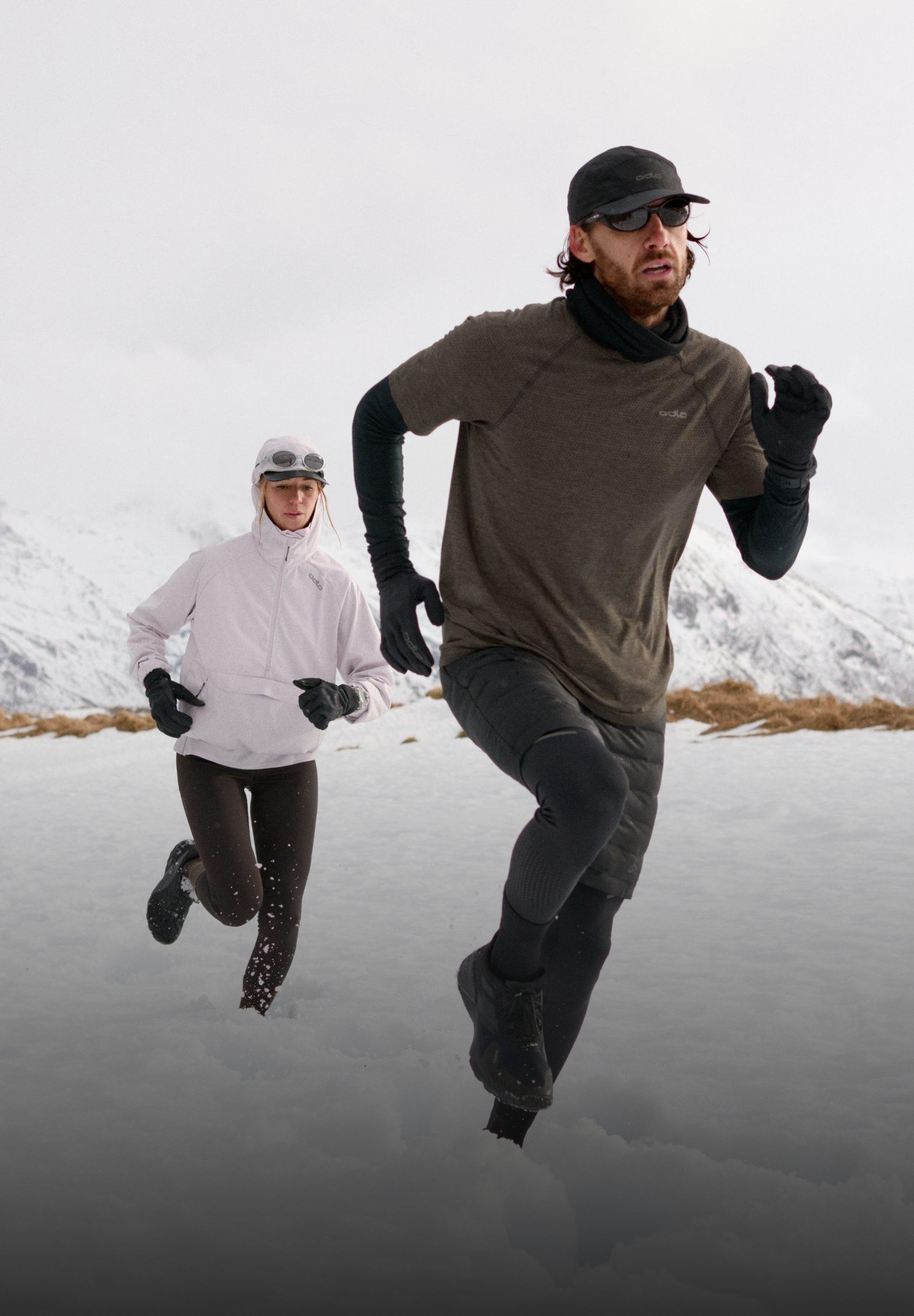 Man in dark gear and woman in a light jacket running through deep snow with snowy mountains in the background.