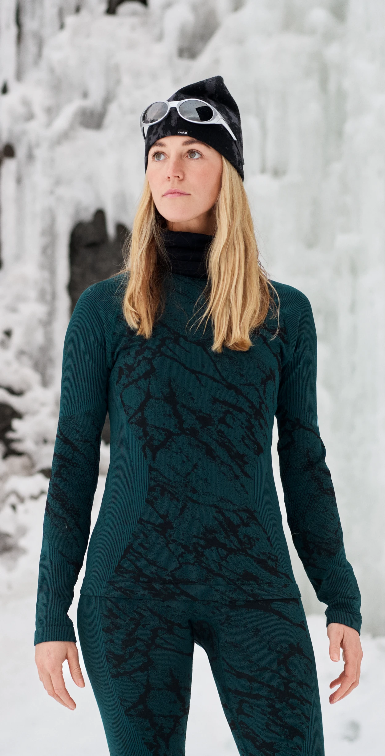 Blonde woman in an icy landscape wearing a dark green and black patterned base layer, black beanie and sunglasses.