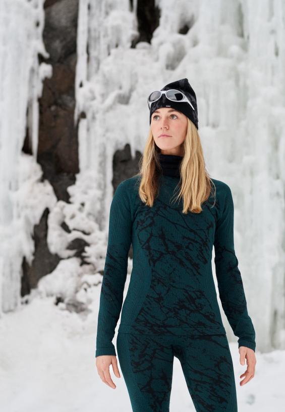 Femme dans un paysage enneigé et glacé portant un sous-vêtement technique à motifs vert foncé et noir, un bonnet noir et des lunettes de soleil.