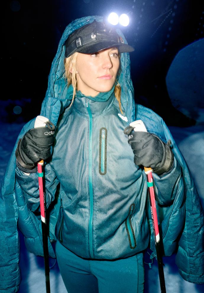 Femme en tenue d'hiver bleu sarcelle et lampe frontale, debout dans la neige de nuit, tenant des bâtons de ski avec une doudoune posée sur la tête.