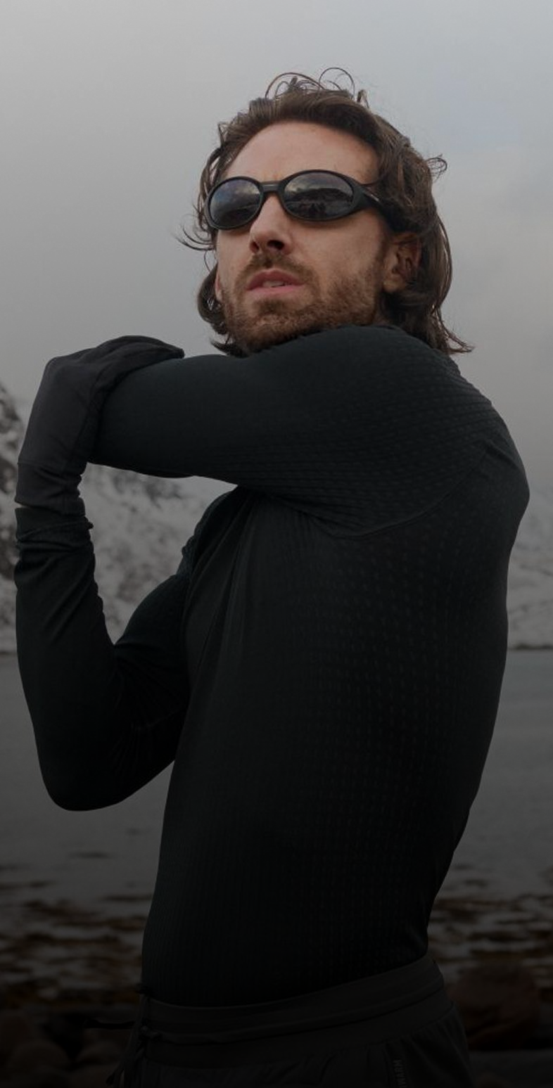 Man in black athletic base layer and sunglasses stretches on a rocky shore by water and snowy mountains.