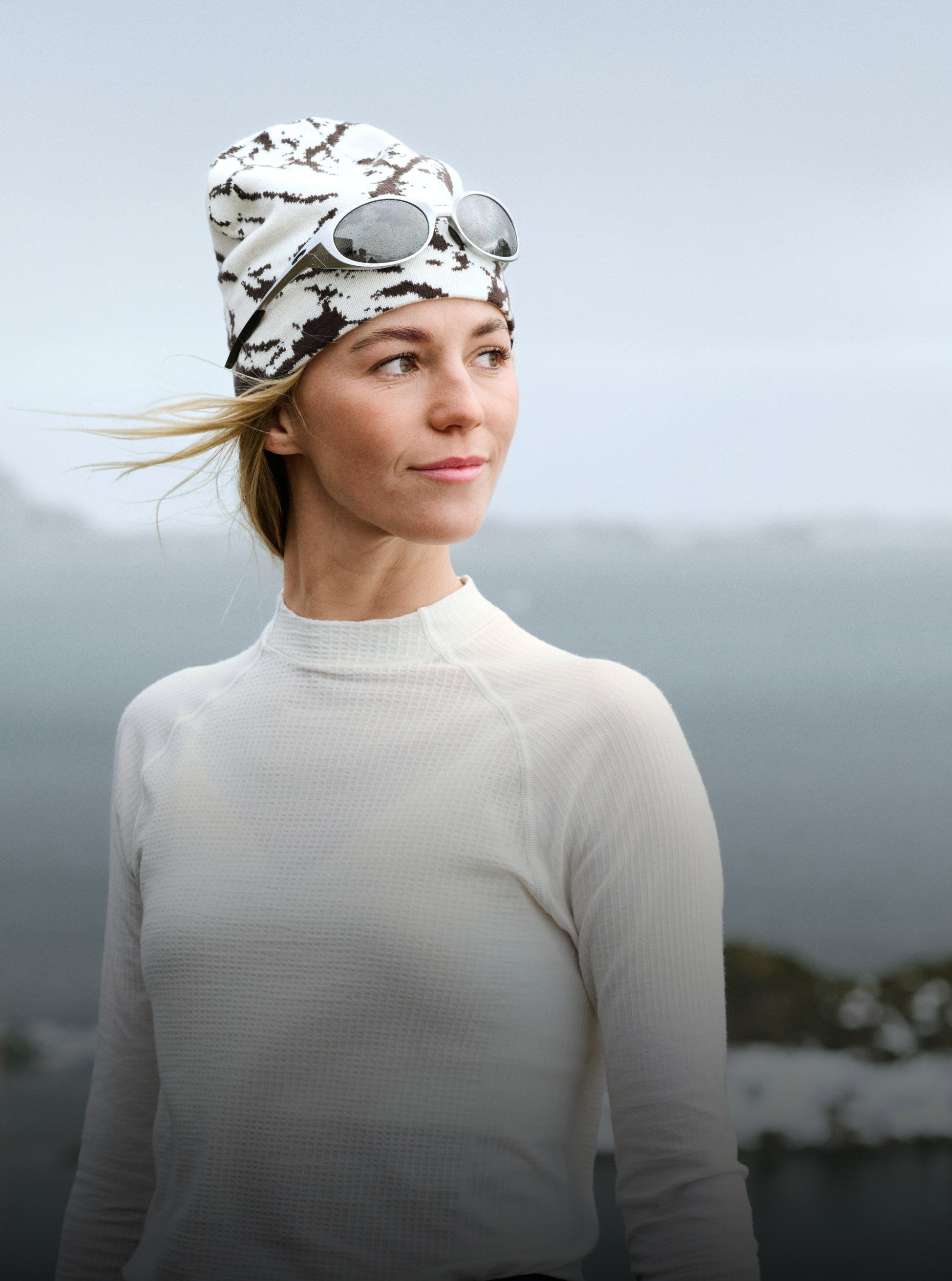 Blonde Frau, die nach rechts schaut, trägt ein weißes Performance Wool Base Layer Oberteil und eine gemusterte Mütze mit Sonnenbrille darauf, vor einem bewölkten grauen Himmel.