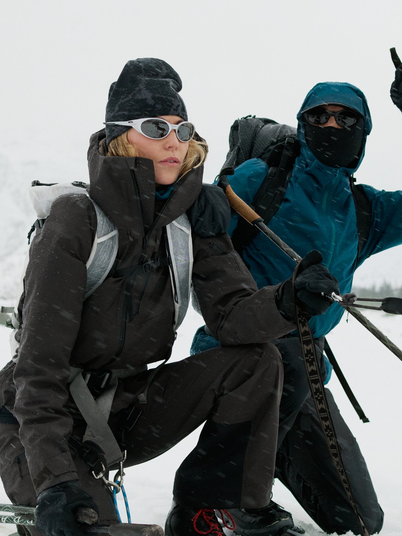 Two people in winter gear, one kneeling in black, one in blue holding poles, with a husky in the snow.
