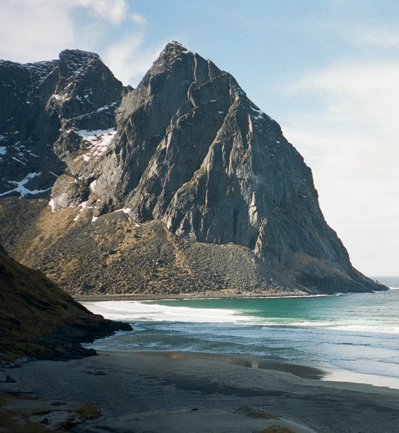 A steep, dark mountain rises beside a sandy beach and turquoise ocean with white waves under a light blue sky.