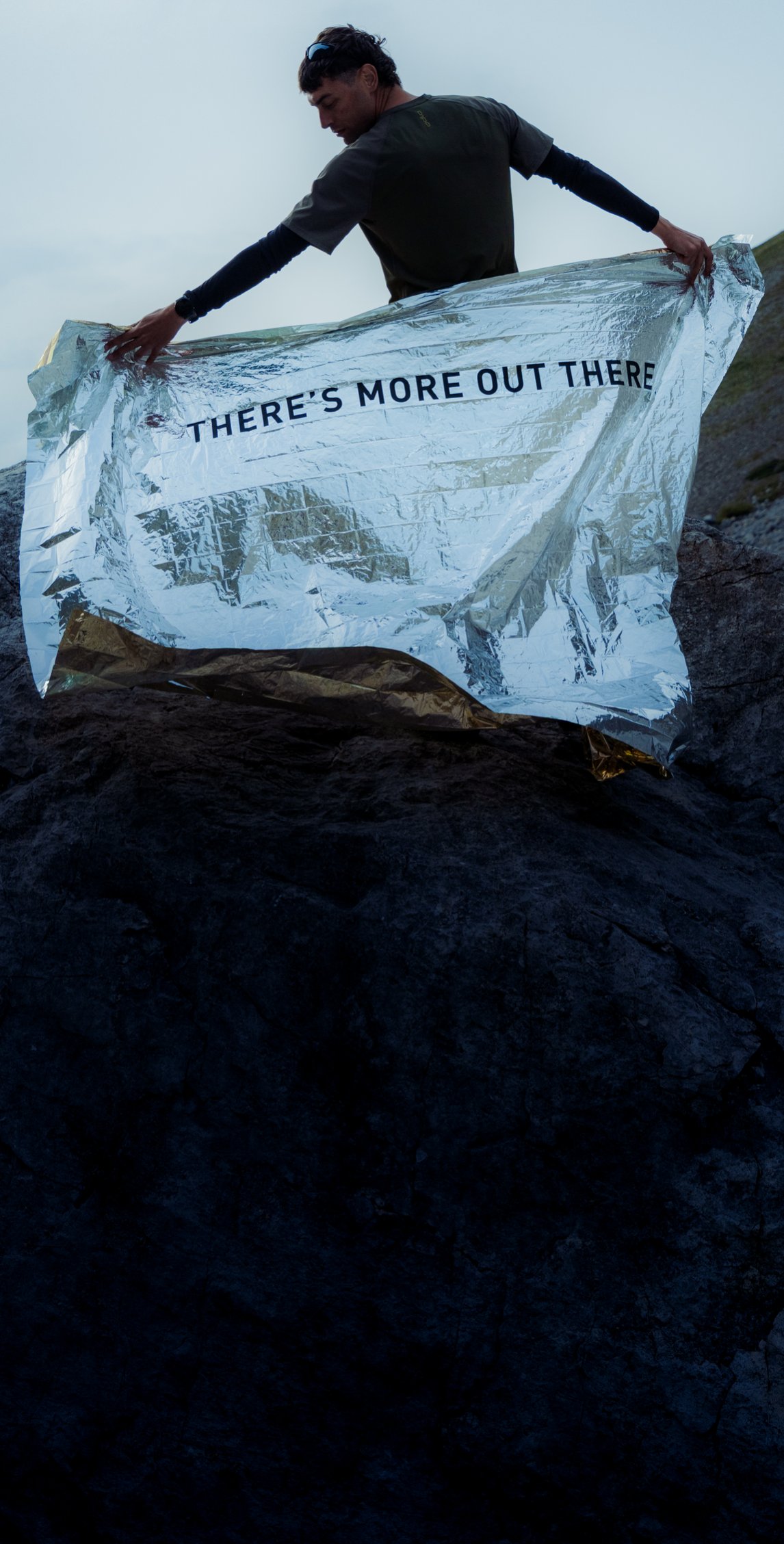 Ein Mann auf einem Felsen in den Bergen hält unter einem bewölkten Himmel eine silberne Folienrettungsdecke mit der Aufschrift „THERE'S MORE OUT THERE“ hoch.