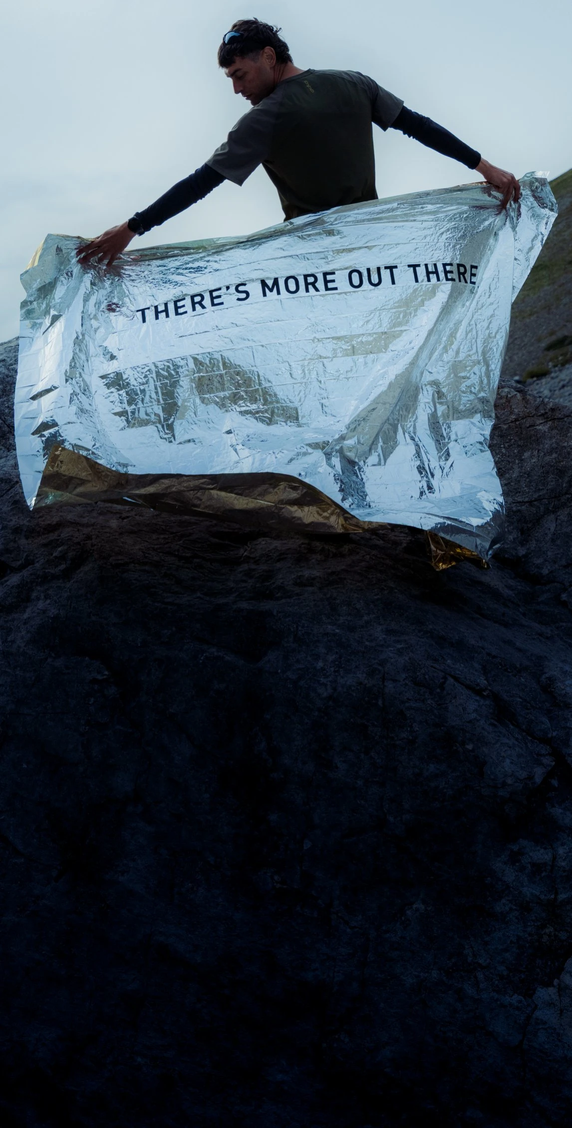 Ein Mann auf einem Felsen in den Bergen hält unter einem bewölkten Himmel eine silberne Folienrettungsdecke mit der Aufschrift „THERE'S MORE OUT THERE“ hoch.
