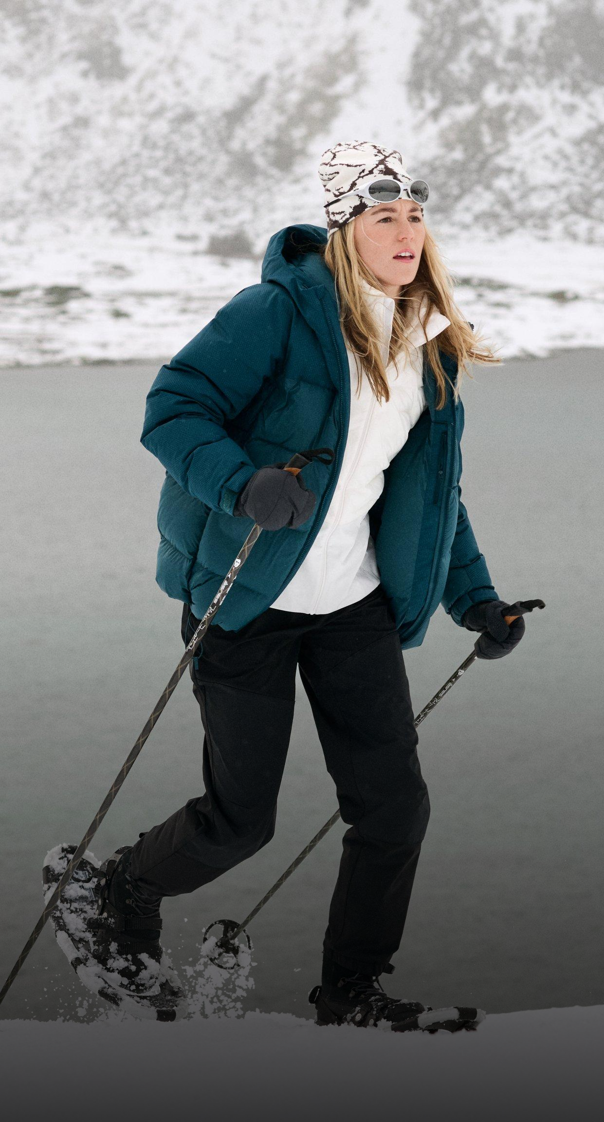 Woman snowshoeing by a snowy lake, wearing a teal puffer jacket, white hat with goggles, and using trekking poles in winter scenery.
