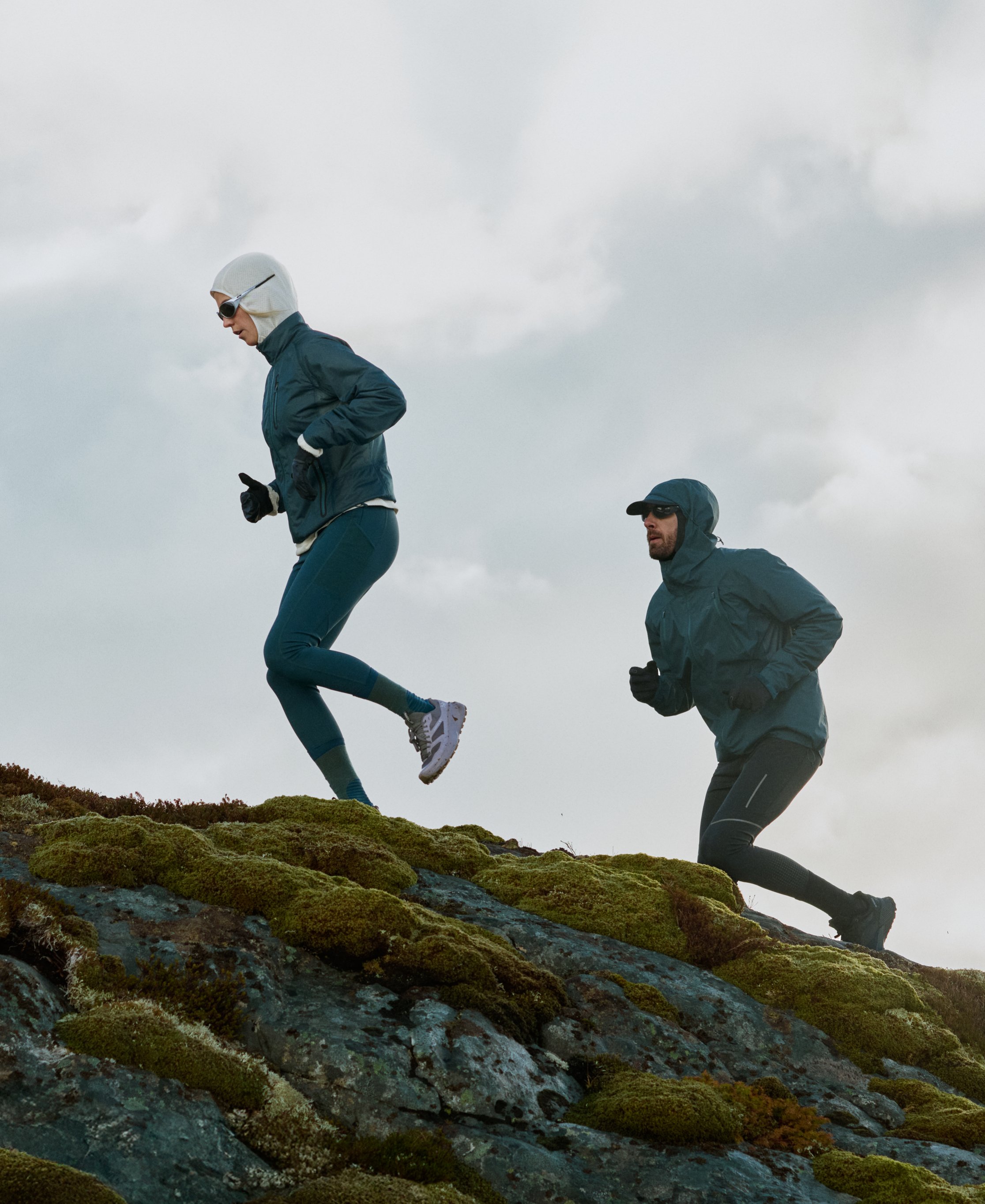 Twee trailrunners in donkerturkooizen jacks en tights rennen een heuvel op over mosbedekte rotsen tegen een bewolkte lucht.