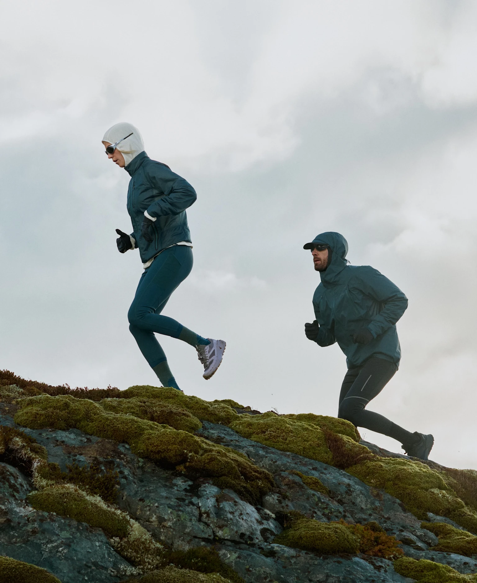 Twee trailrunners in donkerturkooizen jacks en tights rennen een heuvel op over mosbedekte rotsen tegen een bewolkte lucht.