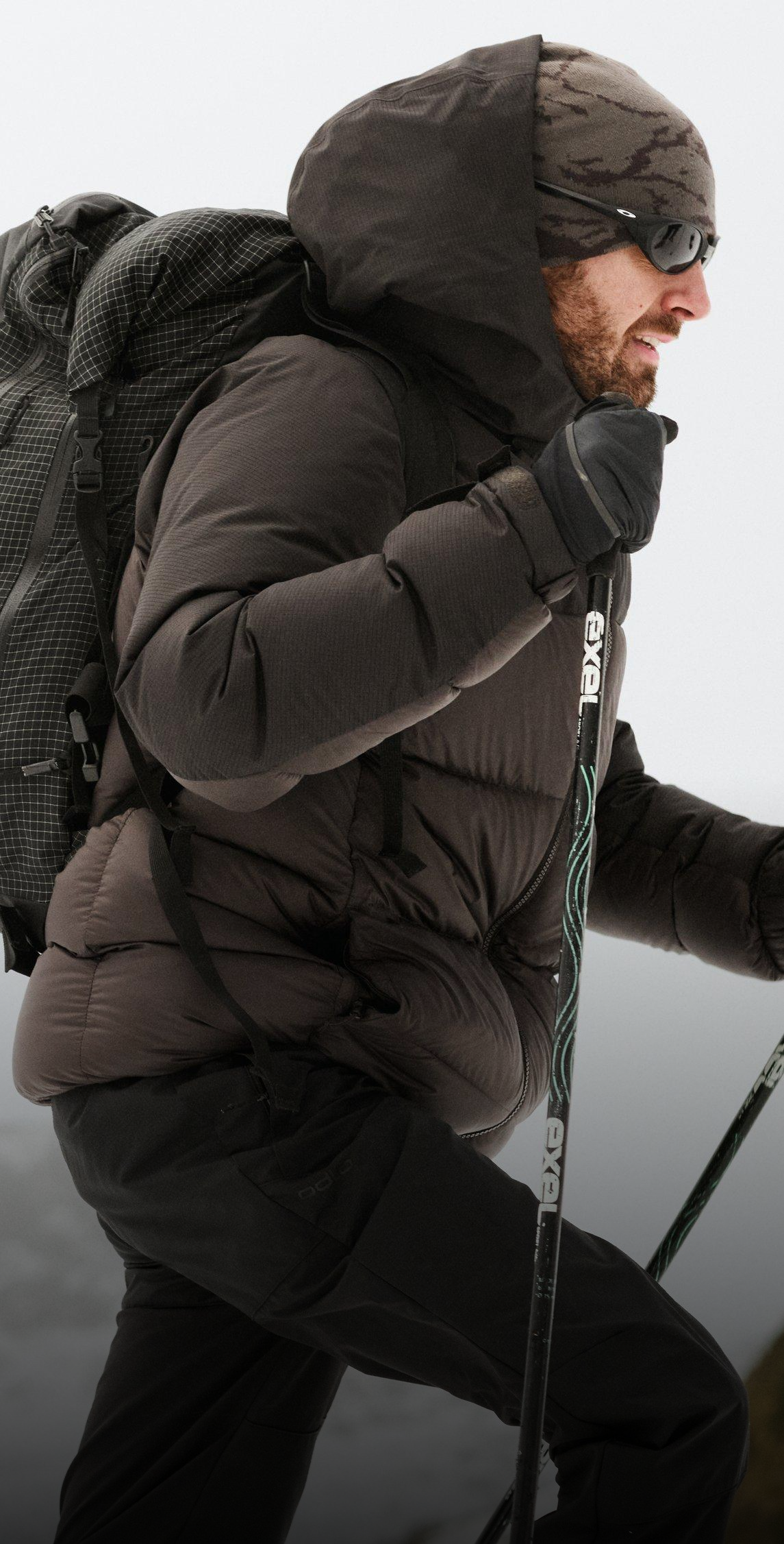 Een man met een baard, in een donkere gewatteerde jas en met een zonnebril, wandelt met een grote rugzak en trekkingstokken door de sneeuw.