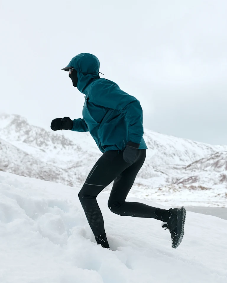 Une personne court sur la neige en montagne, portant une veste bleue à capuche, un pantalon noir et des gants.
