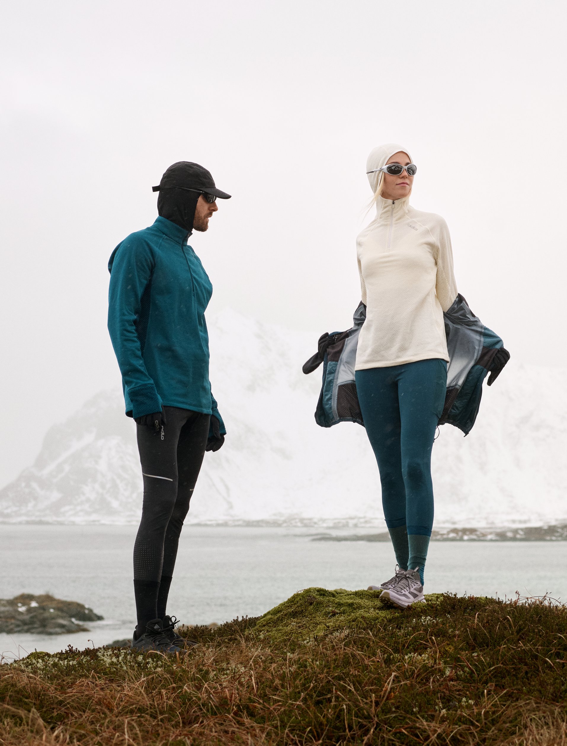 Homme en veste bleue et femme en blanc retirant son manteau sur une colline devant un lac et des montagnes enneigées.