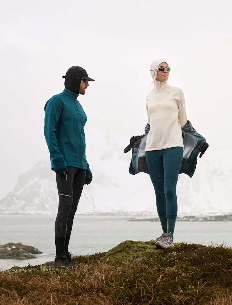 Homme en veste bleue et femme en blanc retirant son manteau sur une colline devant un lac et des montagnes enneigées.