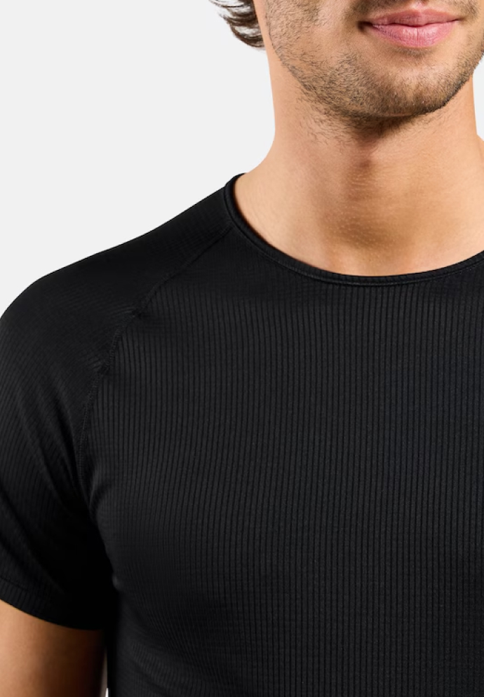 Nahaufnahme eines Mannes in einem eng anliegenden schwarzen, gerippten Kurzarm-Base Layer Shirt, von der Brust bis zum Kinn vor hellgrauem Hintergrund.