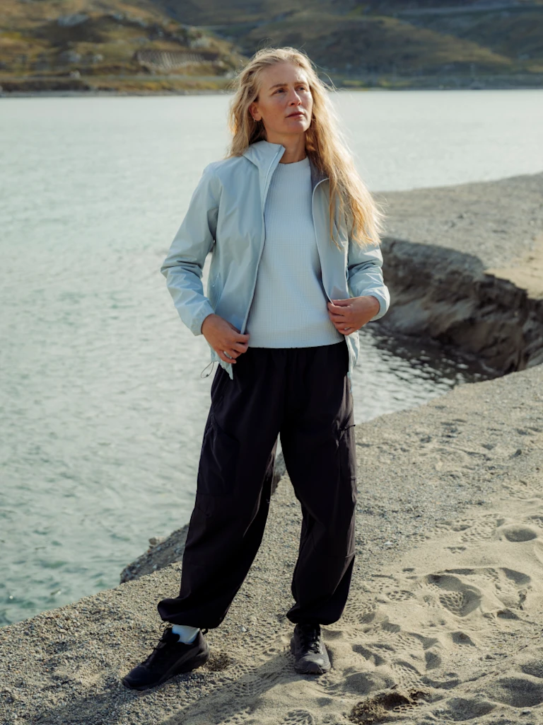 Blonde vrouw bij het water, gekleed in een lichtblauwe jas, licht topje, zwarte broek en sneakers op een zandig strand.