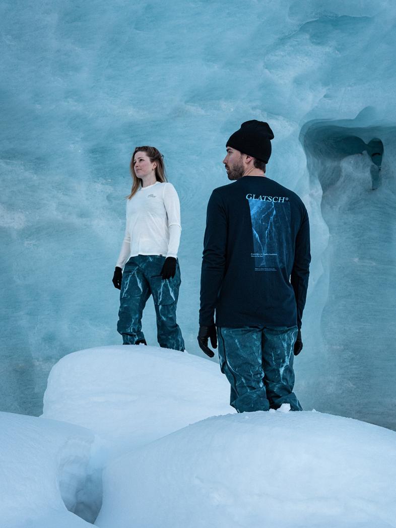 Een man en een vrouw staan op sneeuw in een lichtblauwe ijsgrot. Hij draagt een zwart GLATSCH-shirt; zij draagt een wit topje.