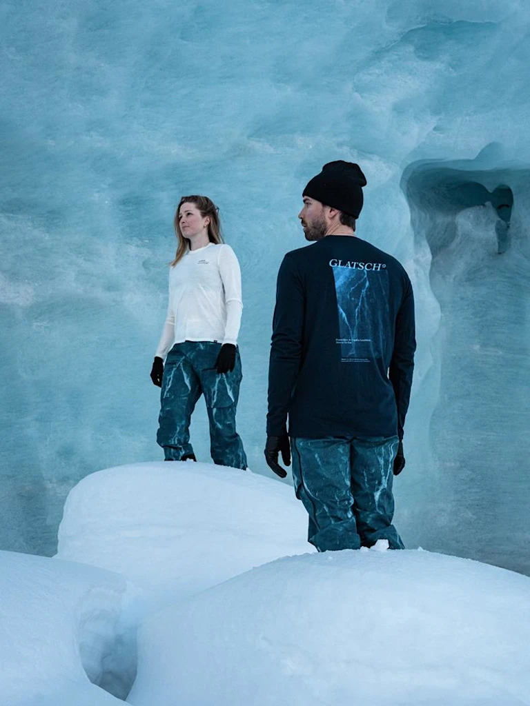 Een man en een vrouw staan op sneeuw in een lichtblauwe ijsgrot. Hij draagt een zwart GLATSCH-shirt; zij draagt een wit topje.