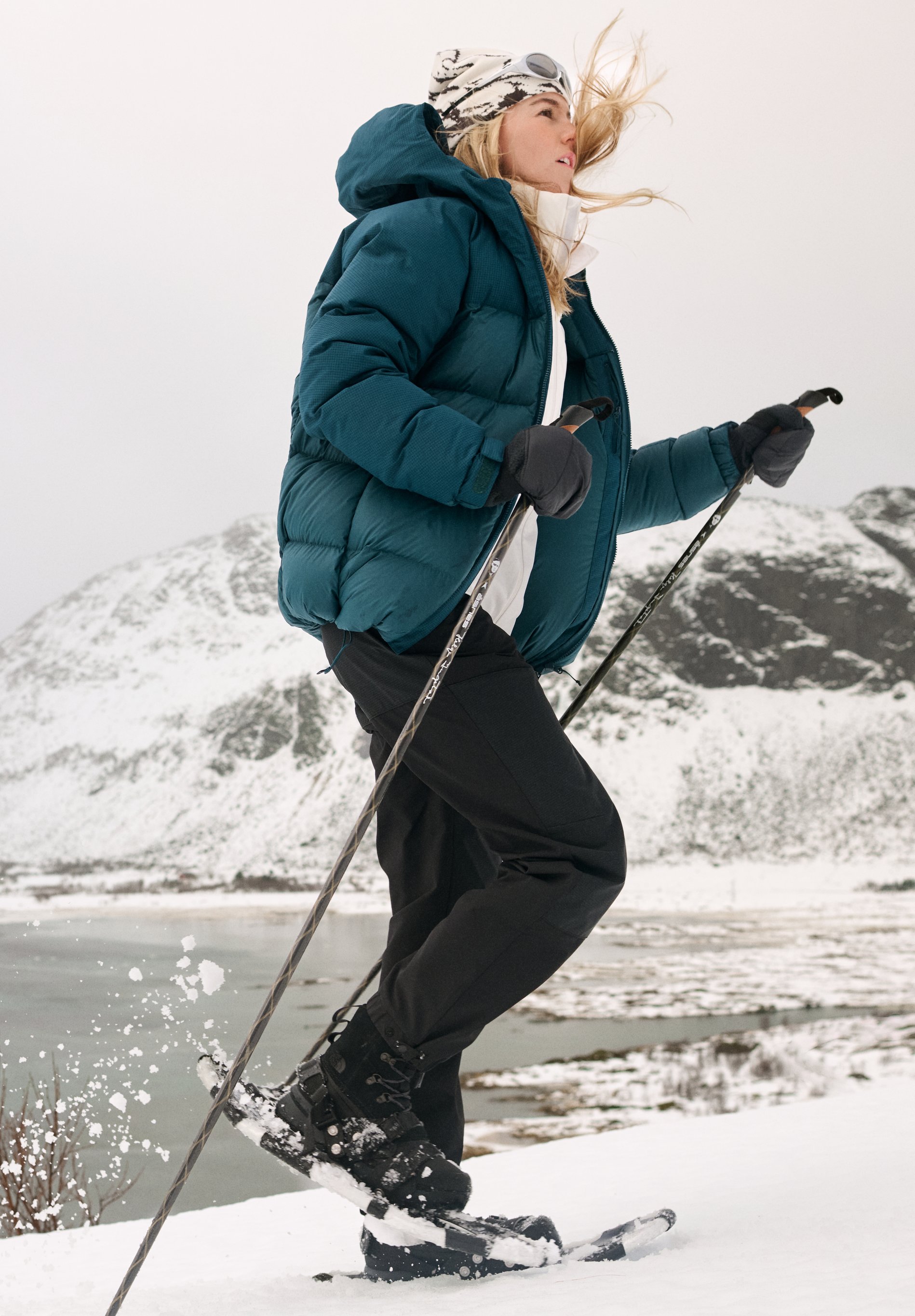 Frau beim Schneeschuhwandern in einer verschneiten Berglandschaft. Sie trägt eine türkisfarbene Steppjacke, eine schwarze Hose und benutzt Skistöcke.