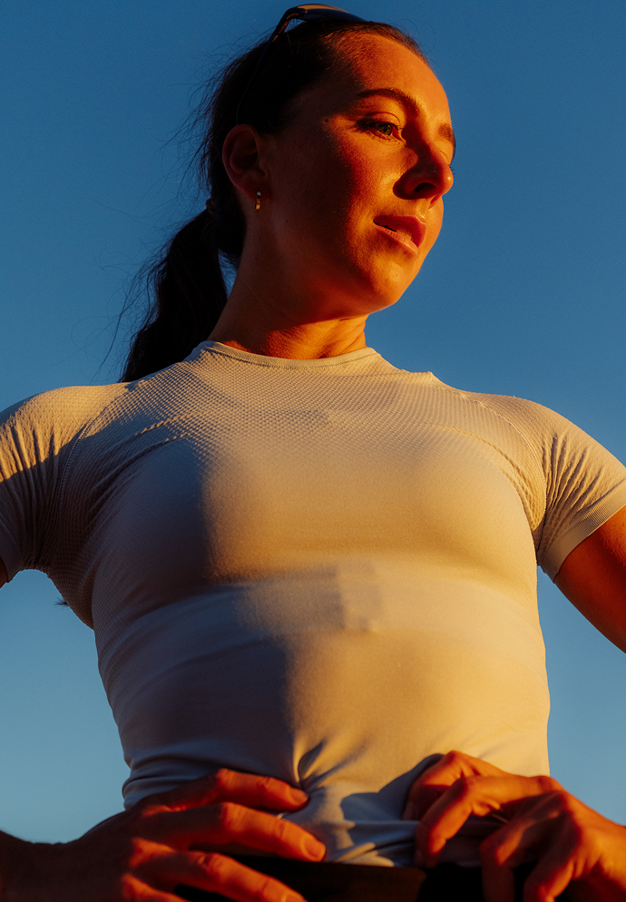 Woman in a light summer base layer standing with hands on hips against a blue sky, illuminated by warm golden hour sunlight.