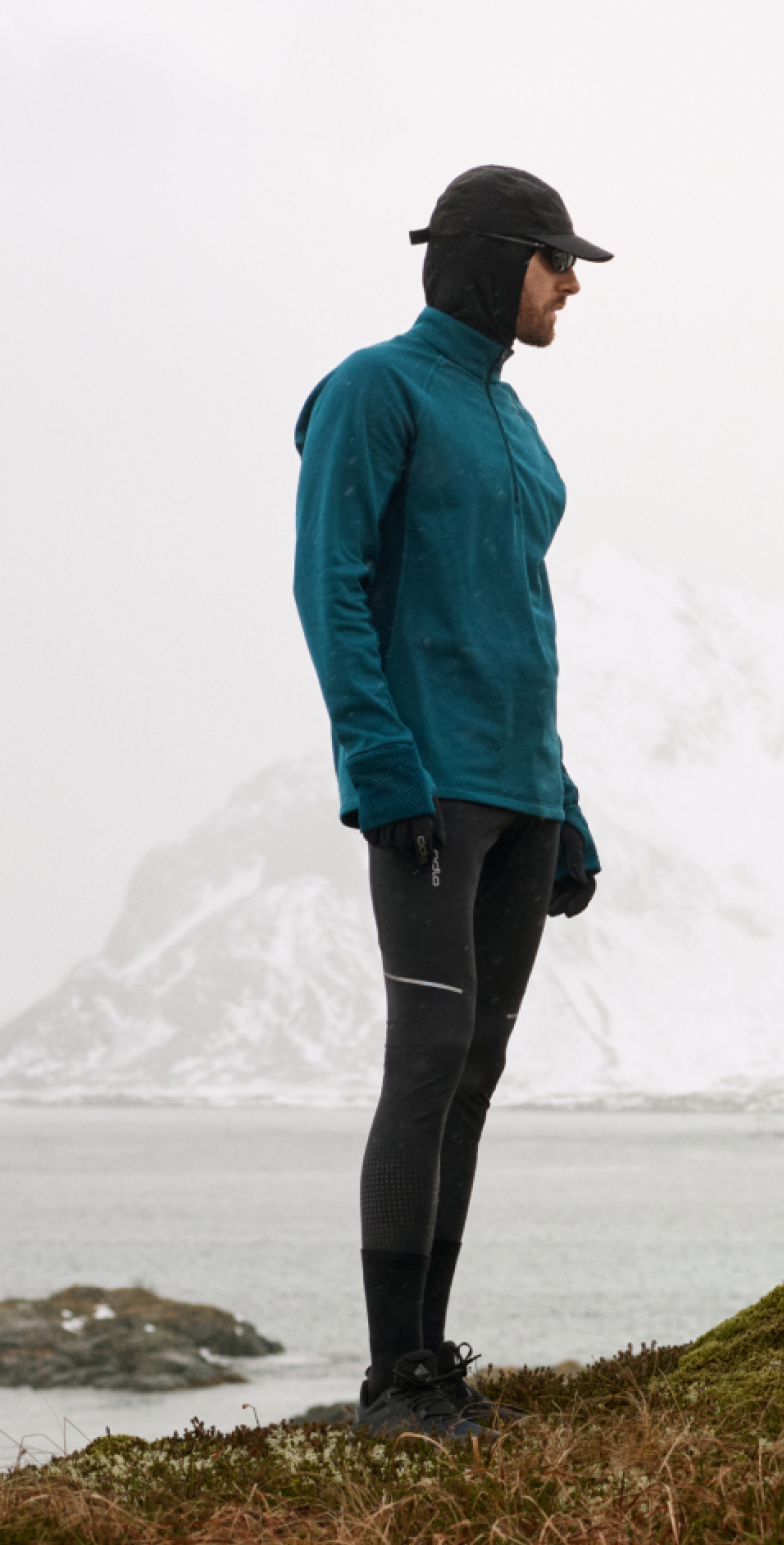 Un uomo con giacca blu e una donna con top bianco sono in piedi su una collina di muschio vicino all'acqua e alle montagne innevate. Lei si sfila un cappotto.