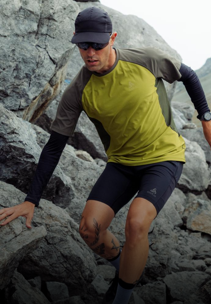A man in a green shirt, black shorts, cap, and sunglasses climbs over large grey rocks in a mountainous area.