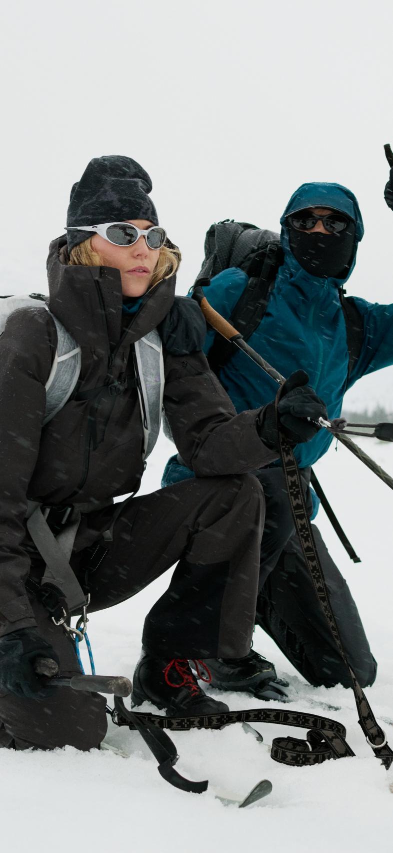 Two people in winter gear kneel in snow. A woman in black with white sunglasses looks ahead; a person in blue points up.