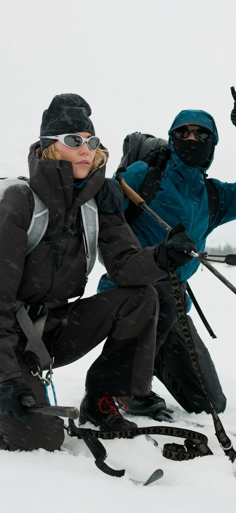 Zwei Personen in Winterbekleidung knien im Schnee. Eine Frau in Schwarz mit weißer Sonnenbrille blickt nach vorne, eine Person in Blau zeigt nach oben.