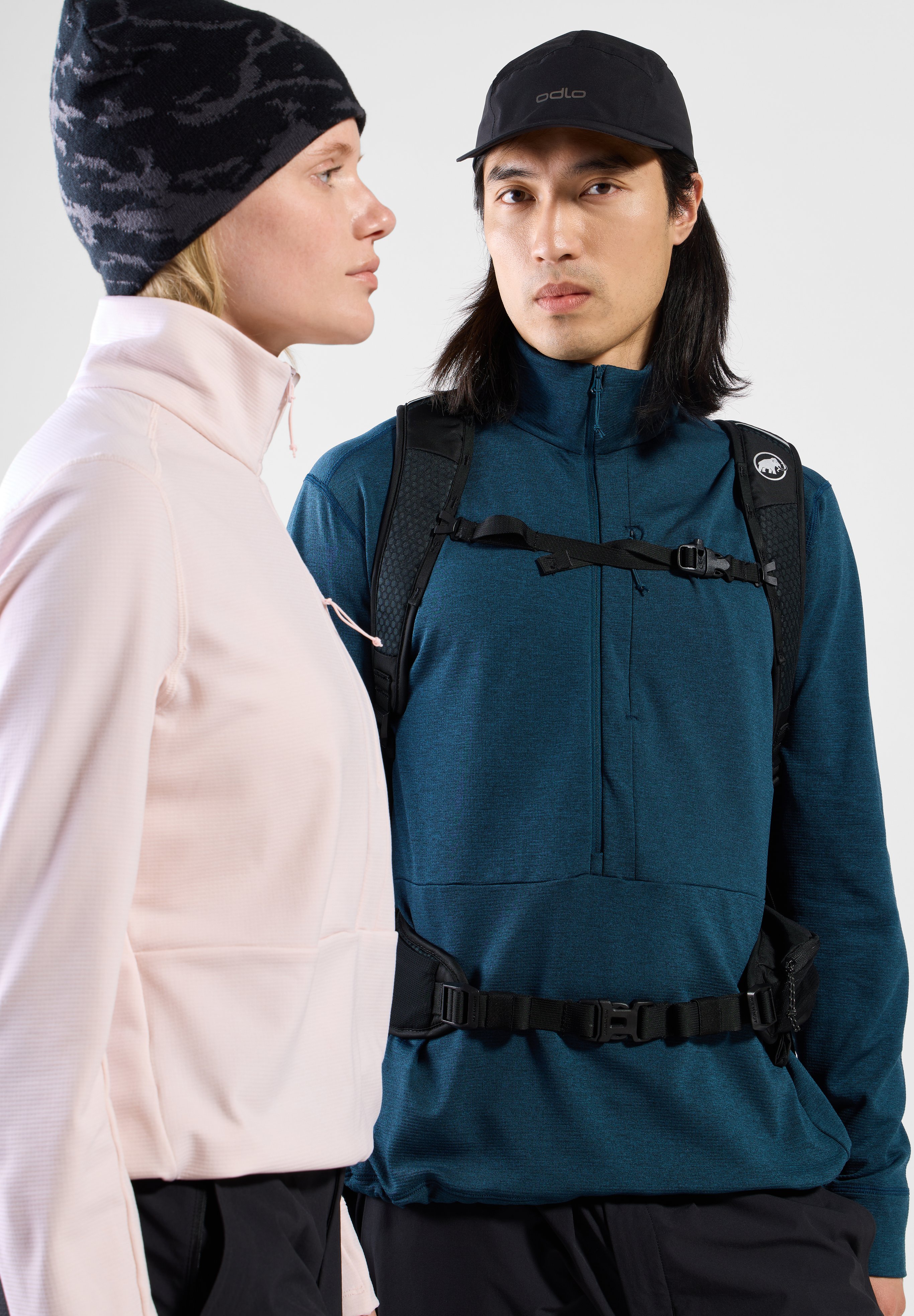 Woman in brown top, beanie and tied jacket, beside a man in a blue hoodie and backpack. Both model outdoor hiking apparel.