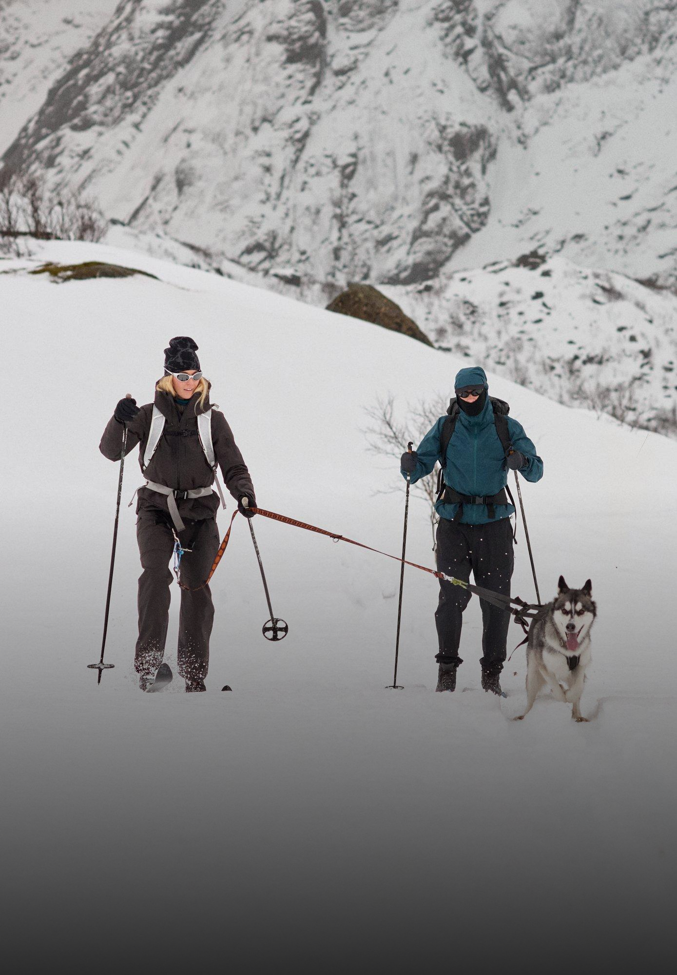 Twee mensen die skiën in een besneeuwd berglandschap. Een husky aan een lijn trekt een vrouw in het zwart voort, naast een persoon in het blauw.