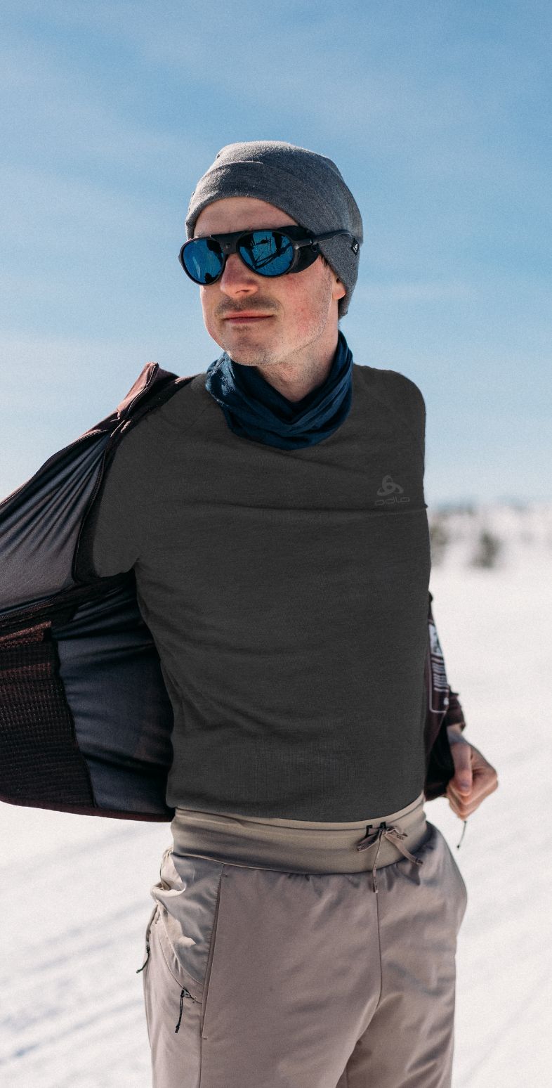 Man in snow opening his jacket to reveal a dark grey base layer, wearing a grey beanie and blue sunglasses.