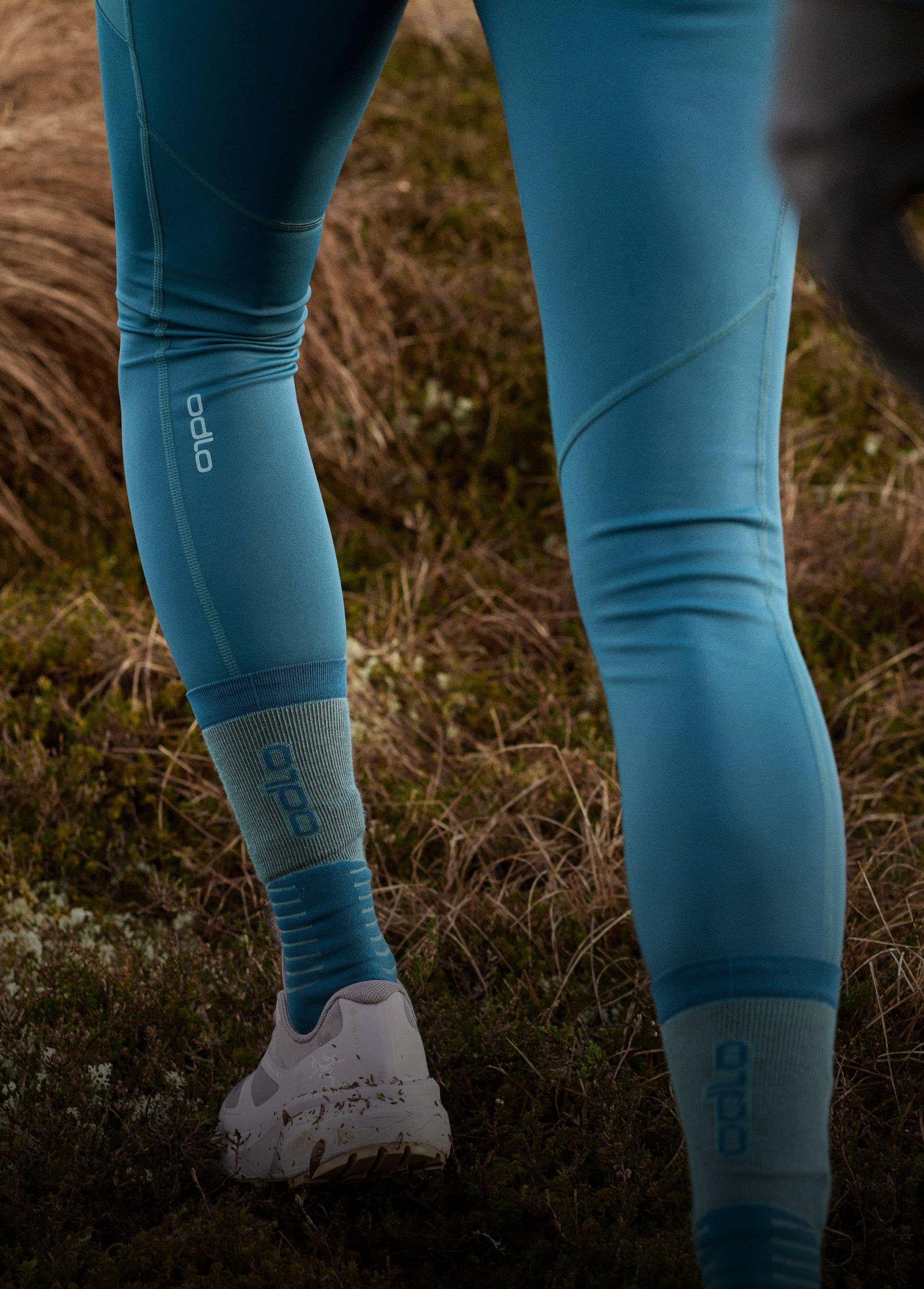 Person trägt türkisfarbene Odlo-Leggings und -Socken mit schlammigen weißen Trailrunning-Schuhen und geht über grasbewachsenes Gelände im Freien.