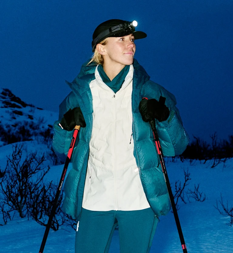 Vrouw in een blauwgroen jack, witte bodywarmer en hoofdlamp staat 's nachts in een besneeuwd landschap met skistokken in de hand.