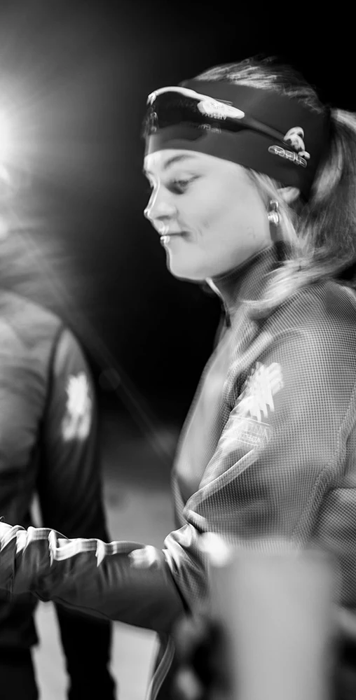 Black and white night photo: A woman pours a steaming drink from a thermos into another's cup. A headlamp shines behind.