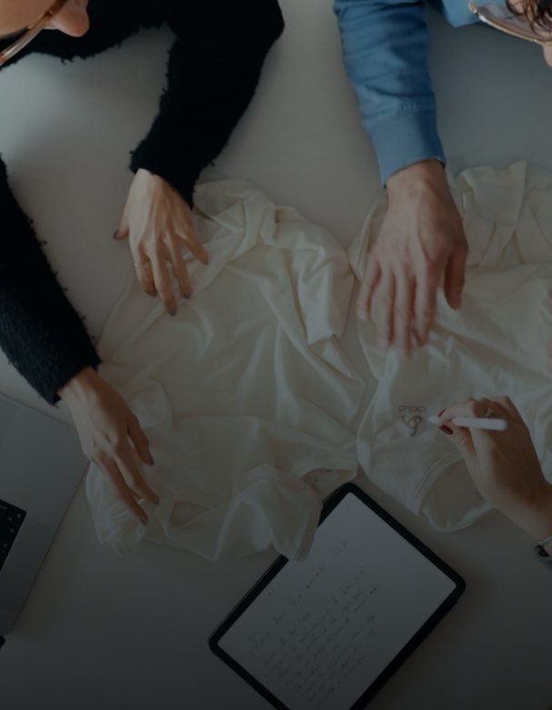 Bovenaanzicht van twee mensen die witte shirts op een bureau bekijken. Een van hen wijst met een stylus naar een logo naast een tablet met notities.